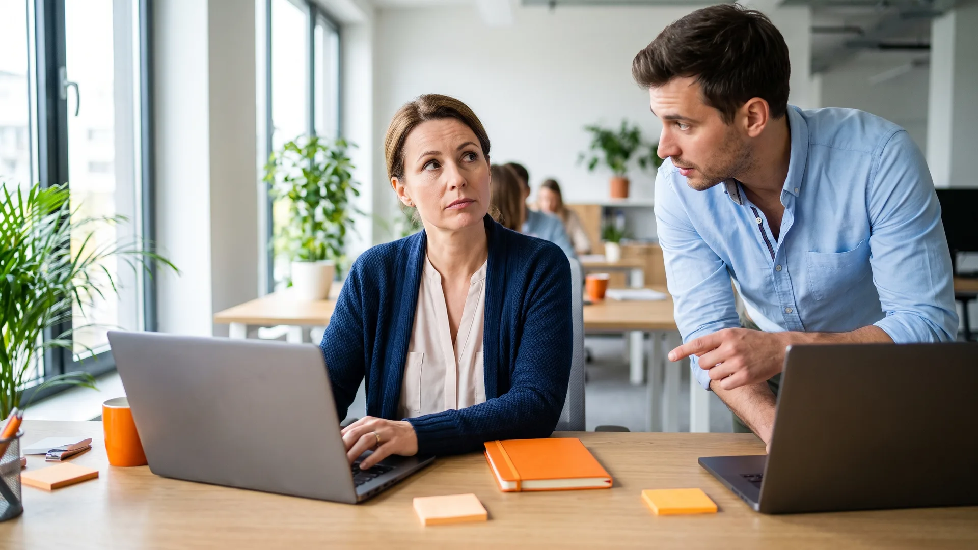 Dans un bureau lumineux, une manageuse interrompt sa relecture sur son ordinateur pour écouter un collègue penché vers elle qui lui montre son propre écran.